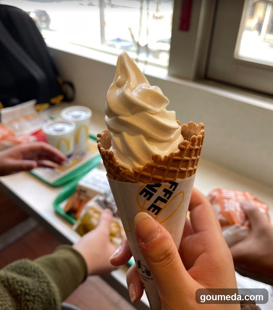 A close-up of a soft-serve ice cream in a waffle cone held by someone inside what appears to be a McDonald’s restaurant. In the background, there are trays with food items and cups on a counter. A person is reaching toward one of the trays. The scene suggests a casual dining environment.