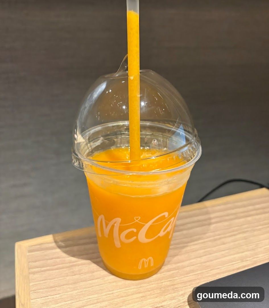 A clear plastic cup filled with bright orange juice, branded with 'McCafé' and the McDonald's logo. The cup has a dome lid with a straw inserted, resting on a light wooden surface.