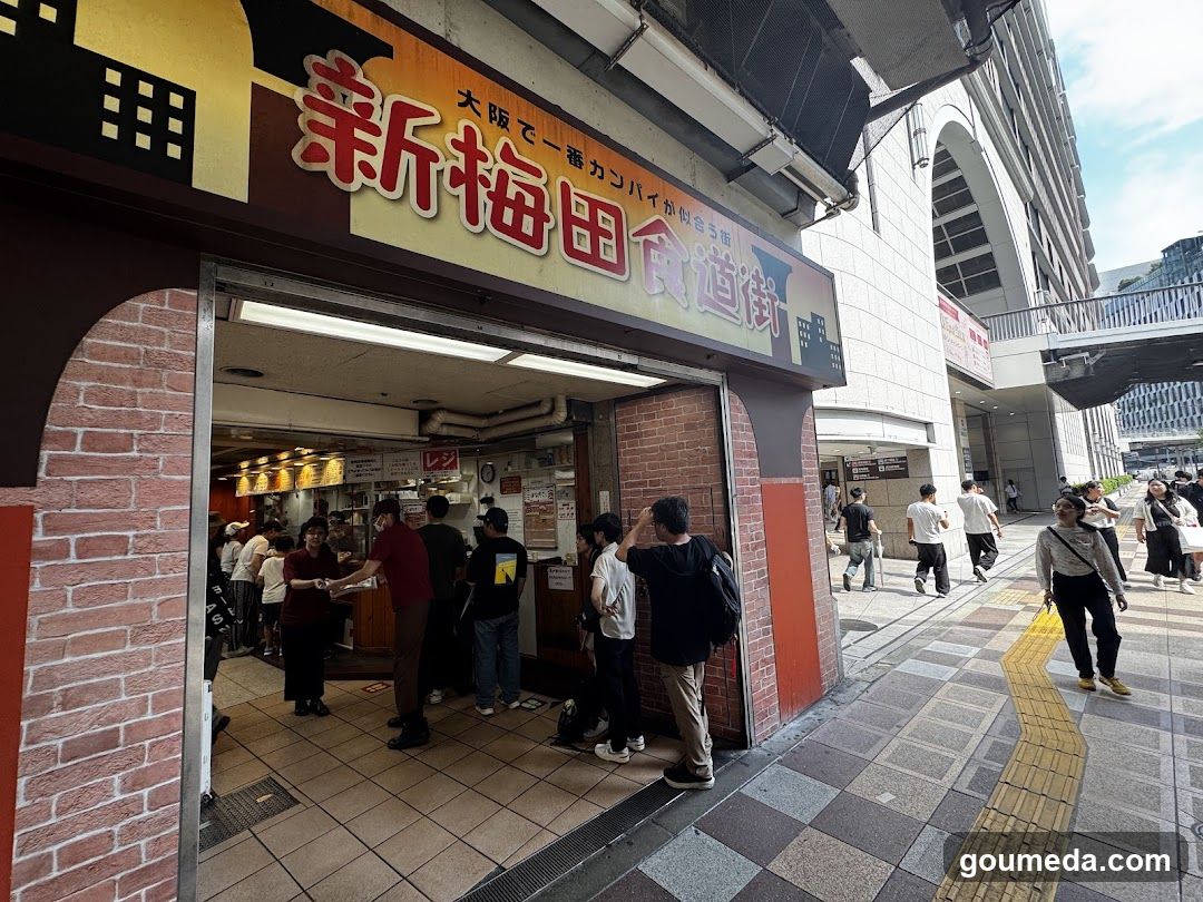 A busy storefront entrance with a large sign reading 'New Ume Street' in Japanese characters, featuring brick columns and people queuing inside.