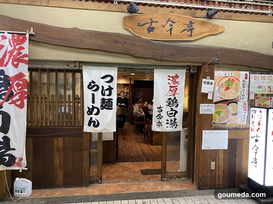 The image shows the exterior entrance of a traditional Japanese ramen shop with wooden signage and banners displaying 'Kokonji' (Ancient Temple) and 'Thick Chicken Soup'. Inside, customers are seated at tables.