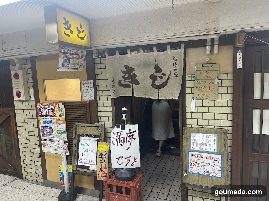 A traditional Japanese restaurant entrance with a noren curtain displaying 'きむ' (Kimu). The facade features yellow brick walls, multiple signs including price lists and advertisements, and a person entering the shop.