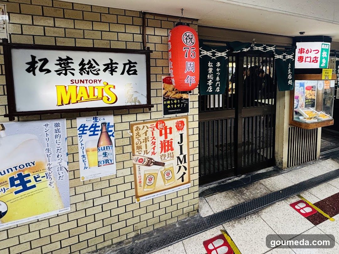 A shop front with a brick facade featuring signage for Suntory Malt's, including a large signboard and various posters advertising malt beverages. A red lantern hangs above the entrance, which is marked by a black gate. There are also signs indicating special promotions and product displays.