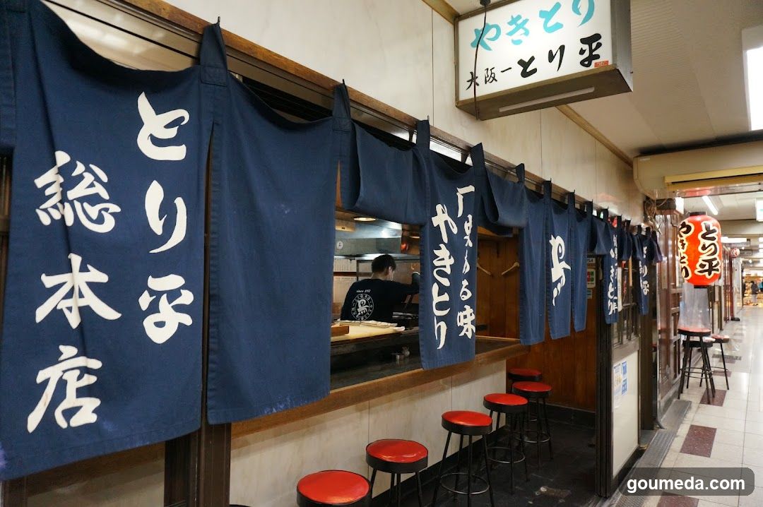 A row of blue noren (door curtains) with white Japanese characters hanging above a counter in what appears to be an indoor market or arcade setting. A sign above reads 'Yaki-Tori Hiro' and 'Osaka - Torihiro'. Red stools are lined up along the counter, and a lantern hangs nearby.