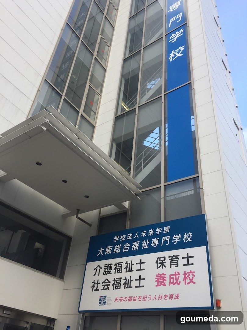 Exterior view of a modern building with glass windows and blue signage displaying Japanese characters. A large signboard at the entrance lists programs such as nursing care worker, caregiver training, social welfare worker, and general education.