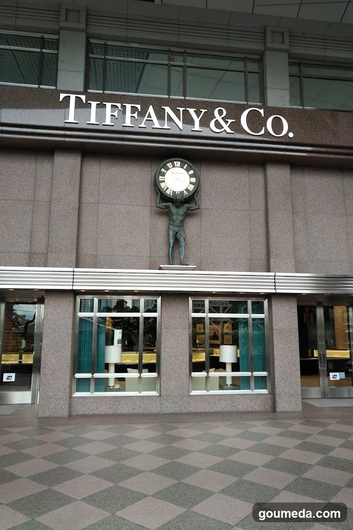 Exterior view of a Tiffany & Co. store featuring the brand's name in white lettering above the entrance. A clock is mounted on the facade, flanked by statues. The ground floor has large glass windows and doors with checkered flooring outside.