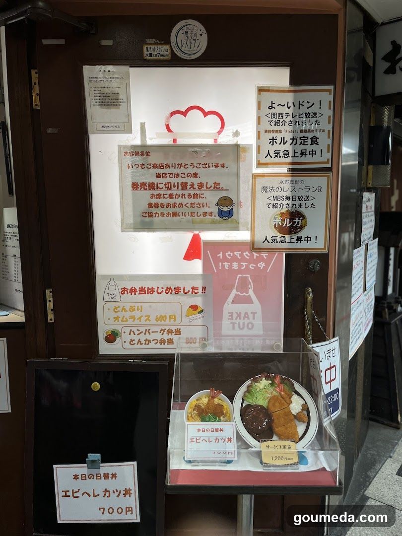 A shop front featuring a wooden door adorned with multiple signs and notices in Japanese. A prominent sign announces 'Take Out' availability, along with other promotional messages about popular dishes like 'Yoi Don'. Below the door is a display case showcasing food items such as 'Ebi Heishikatsu Curry', priced at 700 yen, alongside other menu options.