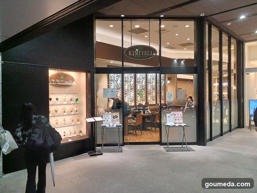 A modern shop entrance with glass doors and windows, featuring a 'KIMIYELL' sign above the door. Inside, there are tables and chairs visible through the glass. Outside, there is a display case on the left showing various products, likely desserts or beverages. A person stands near the display case holding an umbrella.