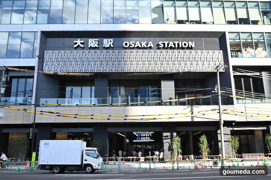 Exterior view of Osaka Station entrance with visible signage reading '大阪駅 OSAKA STATION' on the building facade. A white delivery truck is parked in front, and construction barriers line the sidewalk.