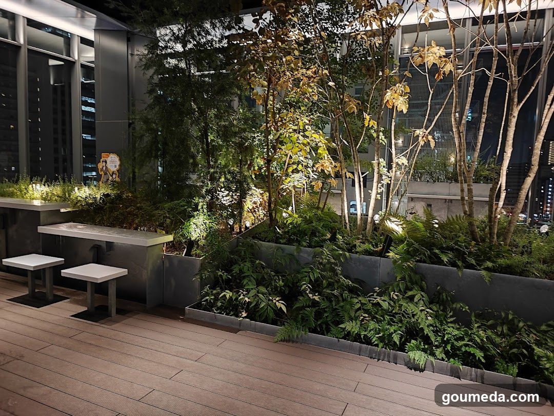 A modern rooftop garden featuring lush greenery, illuminated trees, and minimalist seating on a wooden deck. The scene is set against a backdrop of city lights visible through glass windows.
