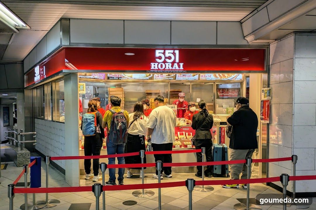 A brightly lit storefront with a red sign displaying '551 HORAI' above the entrance. The shop features glass windows showing food items inside, including stacked boxes and trays of baked goods. Several customers are standing in line outside, separated by red velvet ropes. Staff members wearing masks are visible behind the counter.