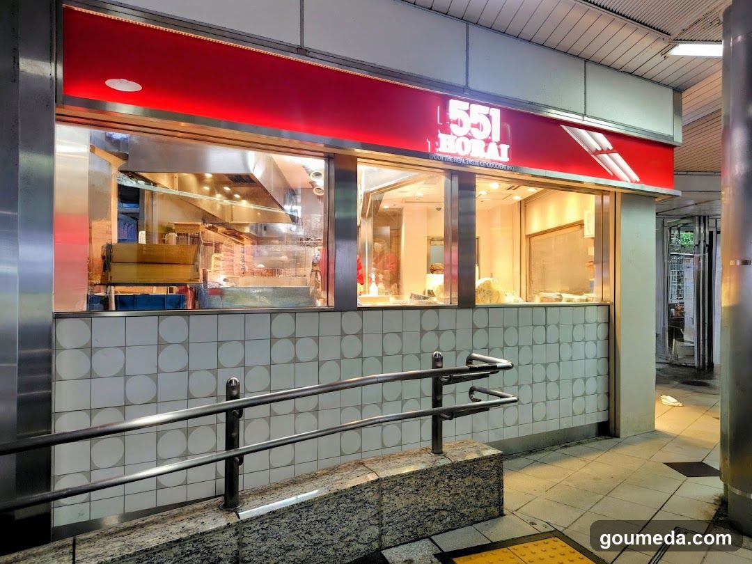 A brightly lit shop front with a red sign displaying '551 MORAI' above large glass windows showing the interior kitchen and food display area. The lower wall is tiled with white square tiles featuring circular patterns. A metal handrail runs along the entrance, leading to a raised granite platform. The scene appears to be in an indoor public space like a mall or transit station.
