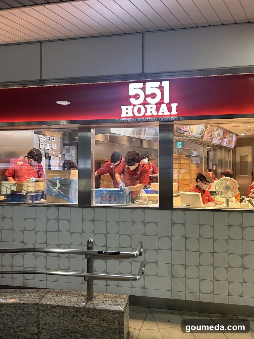 A food stall with a red sign displaying '551 HORAI' and the tagline 'ENJOY THE REAL TASTE OF GOOD EATING'. Staff in red uniforms are visible behind the counter, preparing food. The interior includes stacked boxes and kitchen equipment.