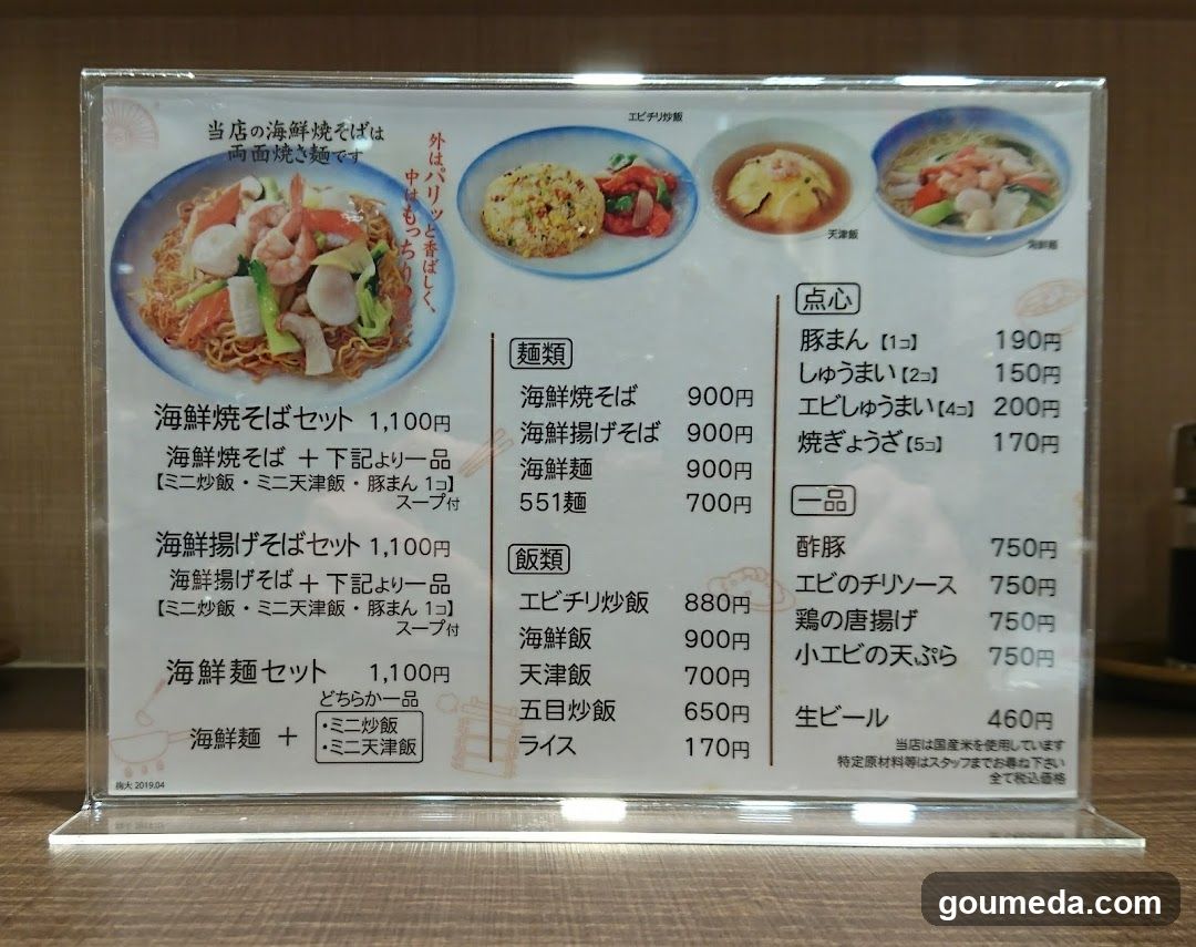 A printed menu board on a clear acrylic stand, displaying seafood soba sets, noodle dishes, rice options, and side items with prices in yen.