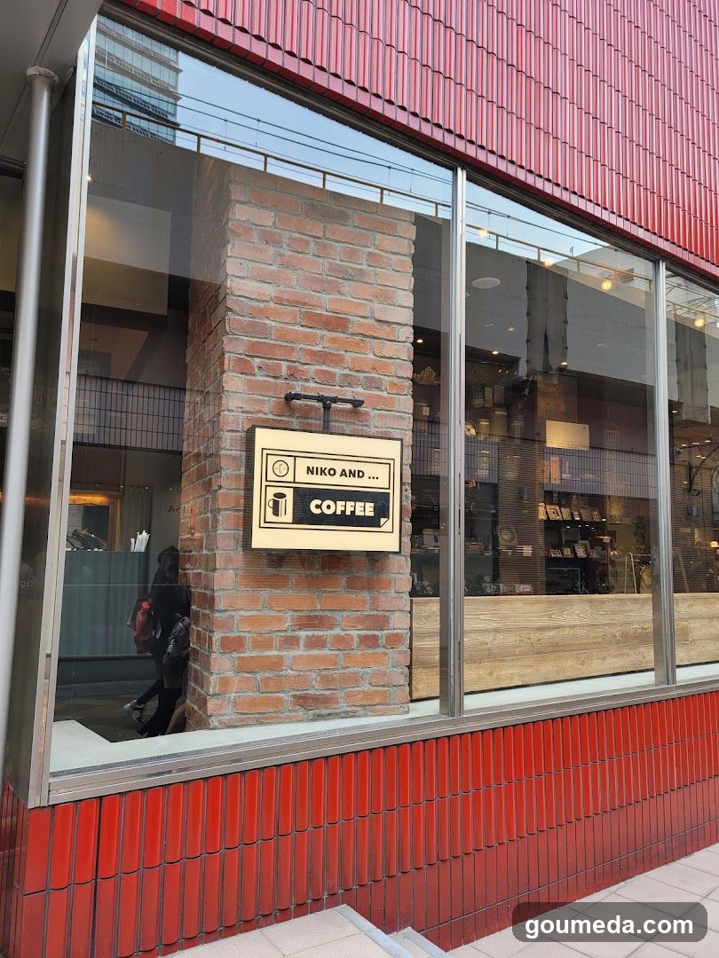 A modern coffee shop exterior featuring a red corrugated metal facade with large glass windows. A brick column in the center displays a sign reading 'NIKO AND ... COFFEE' with icons of a cup and bag. Inside, shelves with products are visible through the glass.