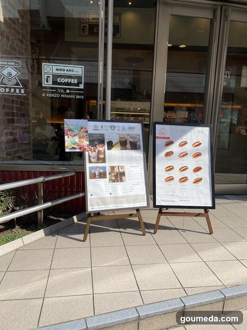 A coffee shop storefront featuring glass doors and windows displaying interior seating. Two A-frame menu boards stand outside on the sidewalk, one listing drinks and desserts, the other showing hot food items like sandwiches or rolls. Signage on the window includes 'NIKO AND COFFEE' and mentions Kenzo Minami.