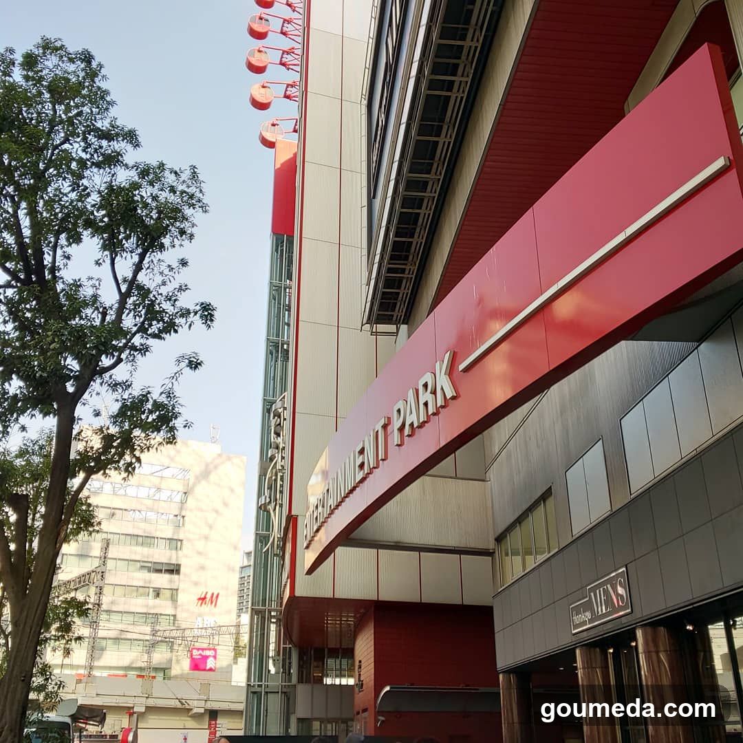 A low-angle shot of the exterior entrance of a modern building with red and white architectural elements. Visible signage includes 'ENTERTAINMENT PARK' on a curved awning, an H&M logo on a distant building, and a 'MENS' sign above glass doors at ground level. A tall tree frames the left side under clear daylight.