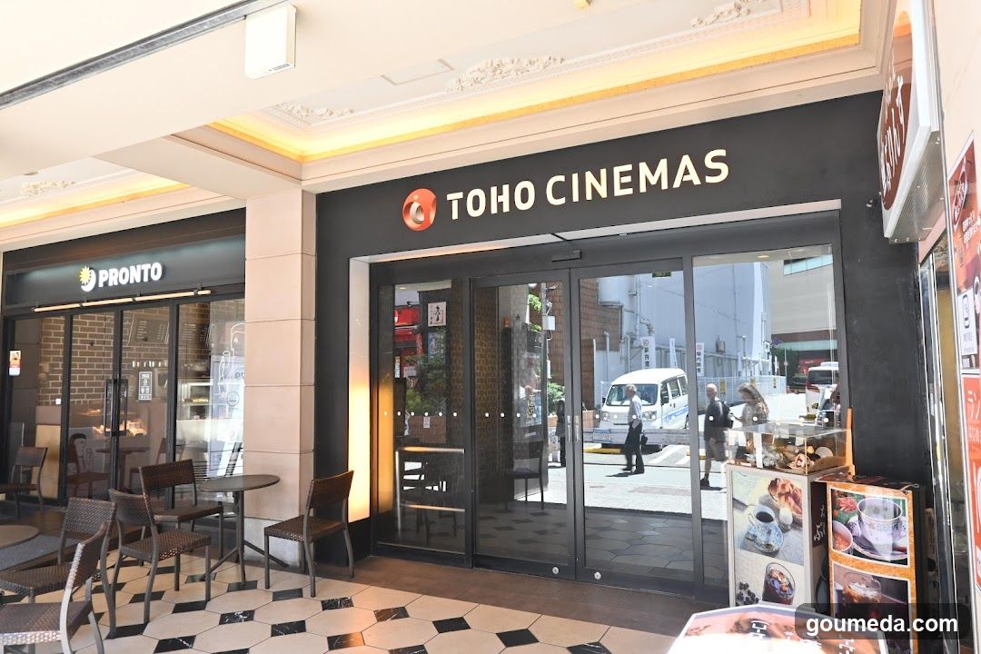 The image shows the exterior of a shopping area with two visible shop fronts. On the left is 'PRONTO', a cafe or restaurant, with outdoor seating and glass doors. To its right is 'TOHO CINEMAS', featuring large glass entrance doors reflecting the street scene outside. A promotional standee displaying food items (coffee, pastries) stands near the cinema entrance. The flooring inside the walkway has black-and-white hexagonal tiles.