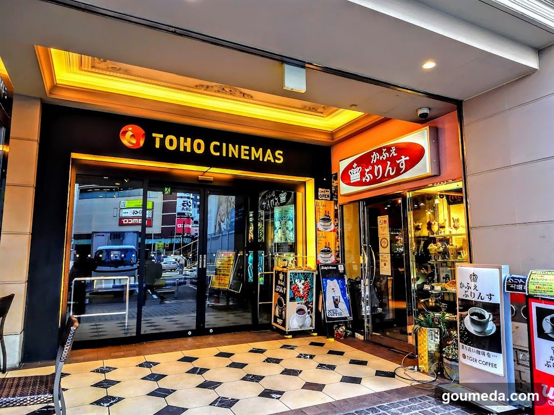 Exterior view of Toho Cinemas entrance with illuminated signage above glass doors. Adjacent is a cafe named 'Kafu no Burinsu' with visible menu boards outside. Checkered floor tiles lead to the entrance.
