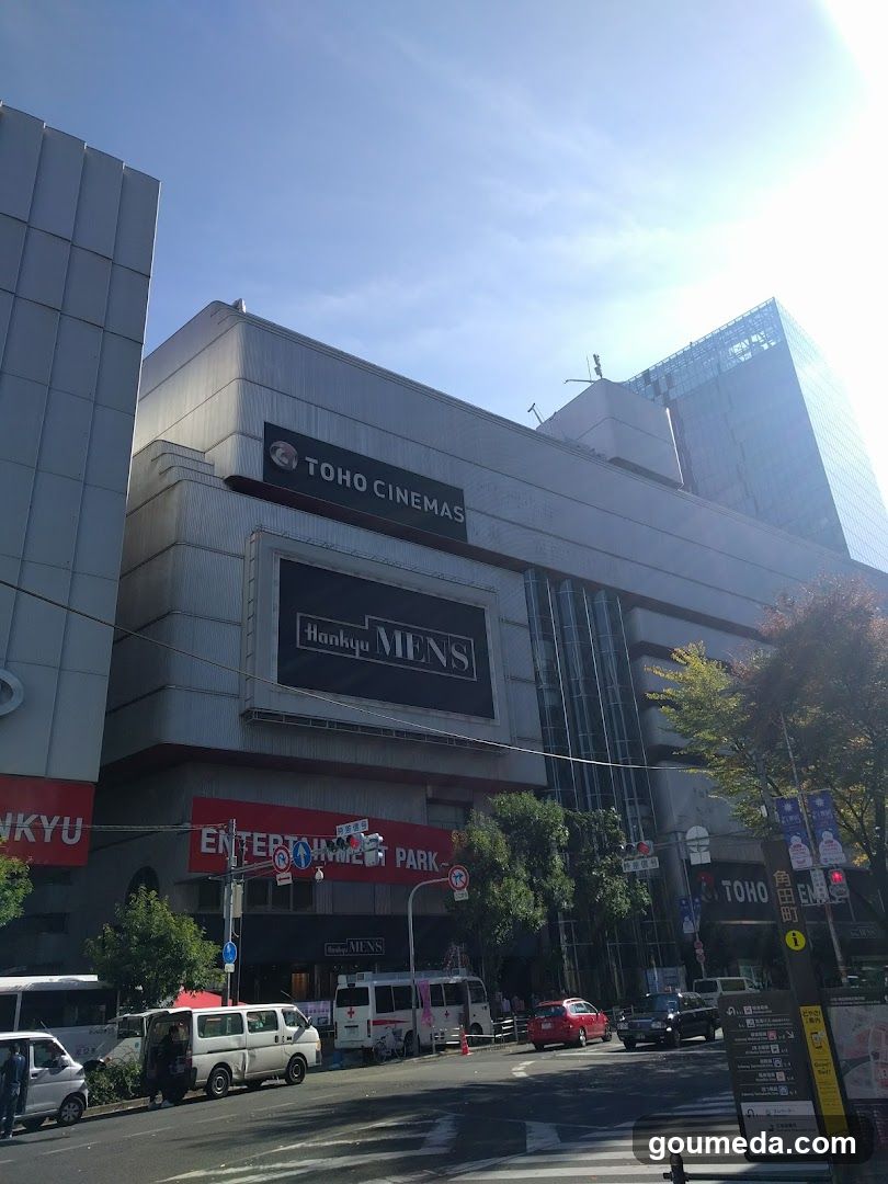 A street-level view of a multi-story commercial building featuring prominent signage for 'TOHO CINEMAS' and 'Hankyu MEN'S'. The facade includes large digital screens, glass windows, and visible entrance areas with vehicles parked nearby under clear skies.