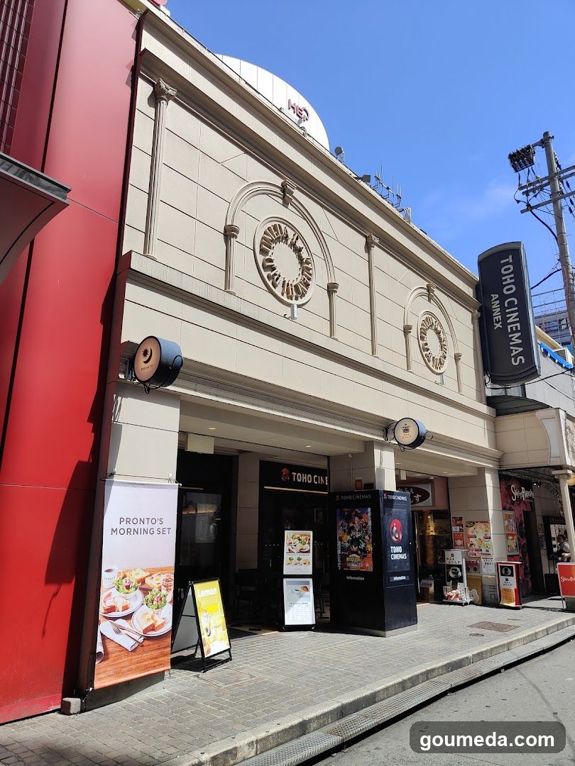 A beige building facade with two circular decorative elements above the entrance. A vertical black sign reads 'TOHO CINEMAS ANNEX'. To the left, a red wall is visible. On the sidewalk, there are promotional posters and signs including one advertising 'PRONTO'S MORNING SET' featuring food images. Another poster displays colorful movie-related graphics.