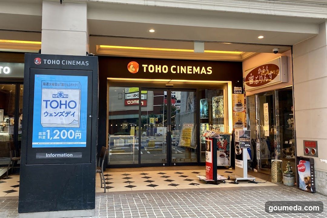 Exterior view of TOHO CINEMAS entrance with illuminated signage, glass doors, and adjacent shop. A digital kiosk displays a promotional ad for 'TOHO Wednesday' at ¥1,200. Adjacent is a small cafe or convenience store with visible menu boards and Coca-Cola branding.