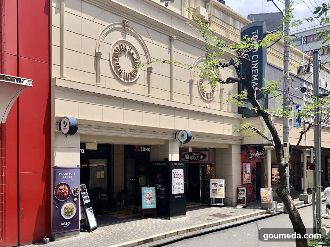 A beige building facade with TOHO CINEMA signage on the right side. The entrance features glass doors and windows, with promotional posters and menu boards outside. A tree branches into the frame from the right.