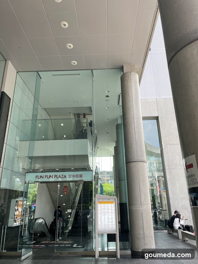 Exterior view of the entrance to Fun Fun Plaza, featuring glass doors and signage with 'FUN FUN PLAZA' and Japanese characters. An escalator is visible inside through the transparent facade.