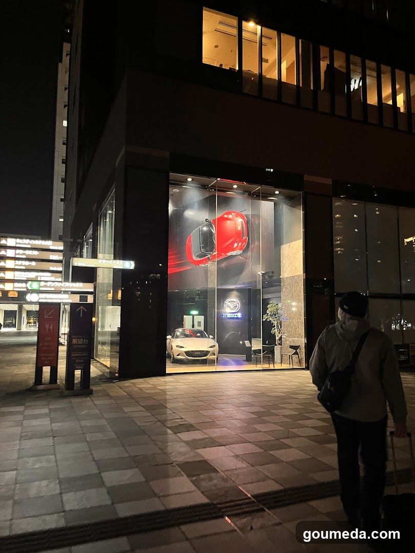 Night view of a Mazda dealership storefront featuring illuminated glass windows displaying red and white cars. A pedestrian walks past the entrance with luggage, while signage points to nearby locations.