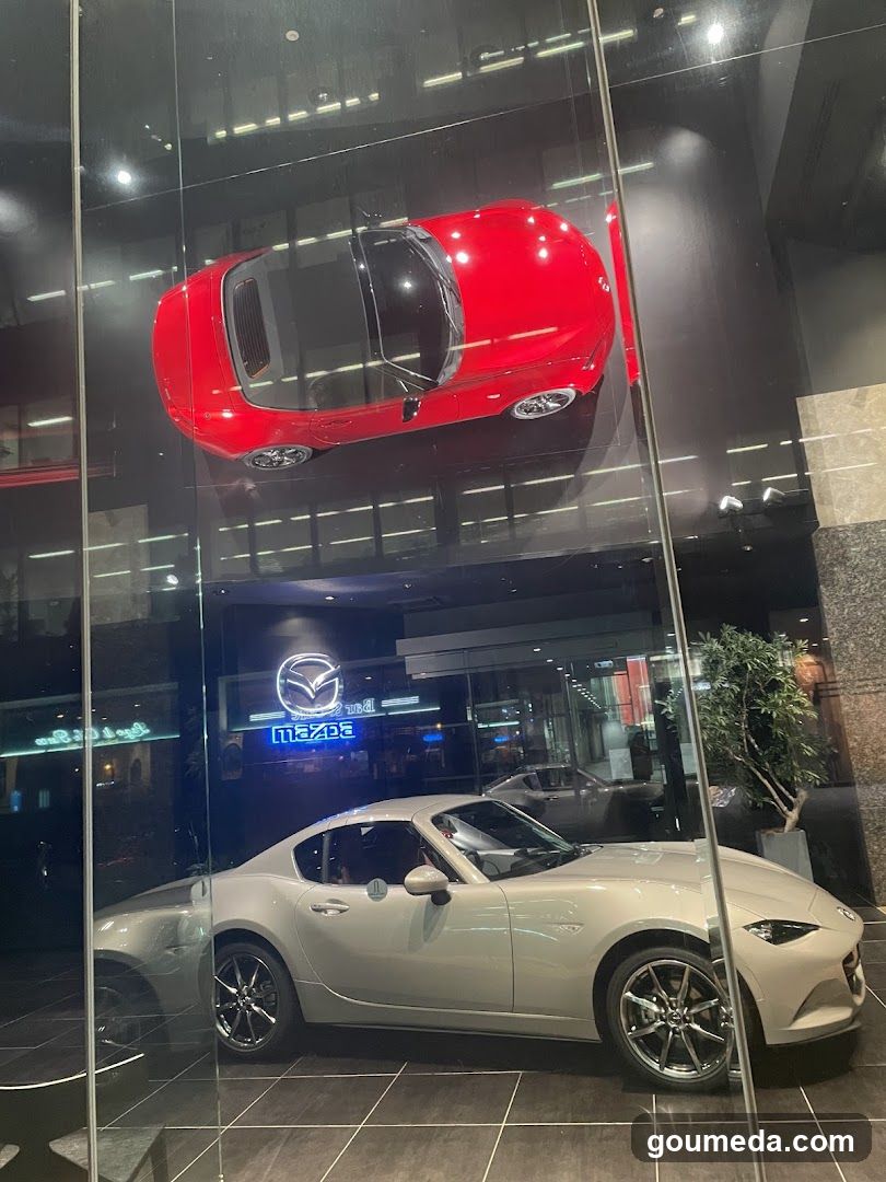 Night view of a Mazda dealership storefront with illuminated signage displaying the Mazda logo and brand name. A white sports car is parked in front, while a red convertible is displayed inside through glass windows above. The building facade features modern lighting and reflections on surrounding surfaces.