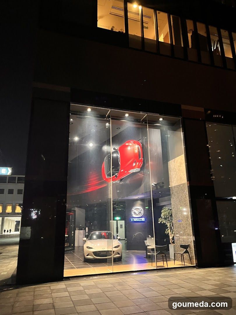 Night view of a Mazda dealership storefront featuring large glass windows displaying a white car and a red vehicle graphic. The building has illuminated signage with the Mazda logo visible inside. The exterior is modern with dark cladding and tiled pavement in front.