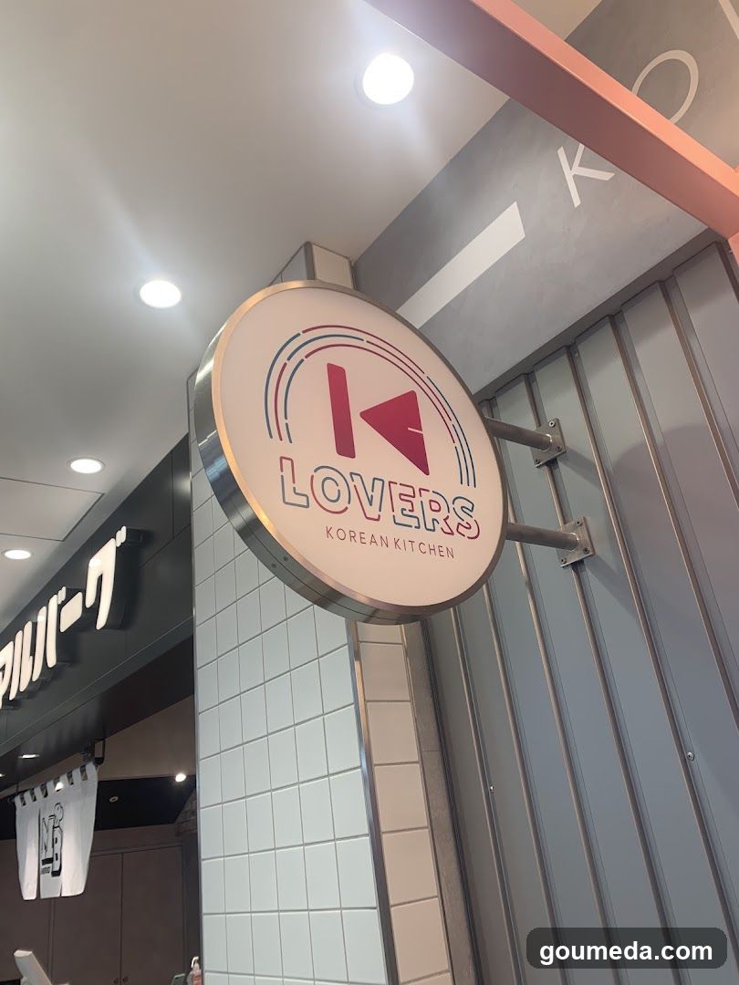 A circular sign with the word 'LOVERS' and a stylized arrow logo is mounted on the exterior wall of a building. The sign also reads 'KOREAN KITCHEN'. To the left, part of another storefront with Japanese characters is visible.