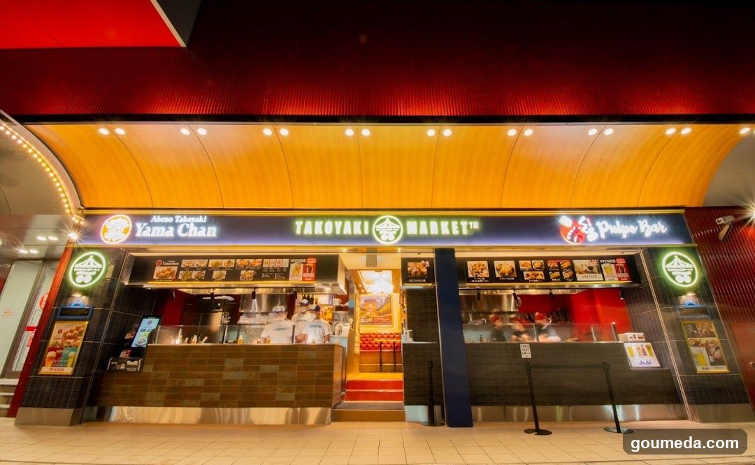 Night exterior shot of a brightly lit food stall complex featuring 'Yama Chan' and 'Takoyaki Market'. The facade has illuminated signage, glass windows showing kitchen activity, and promotional posters on the sides.