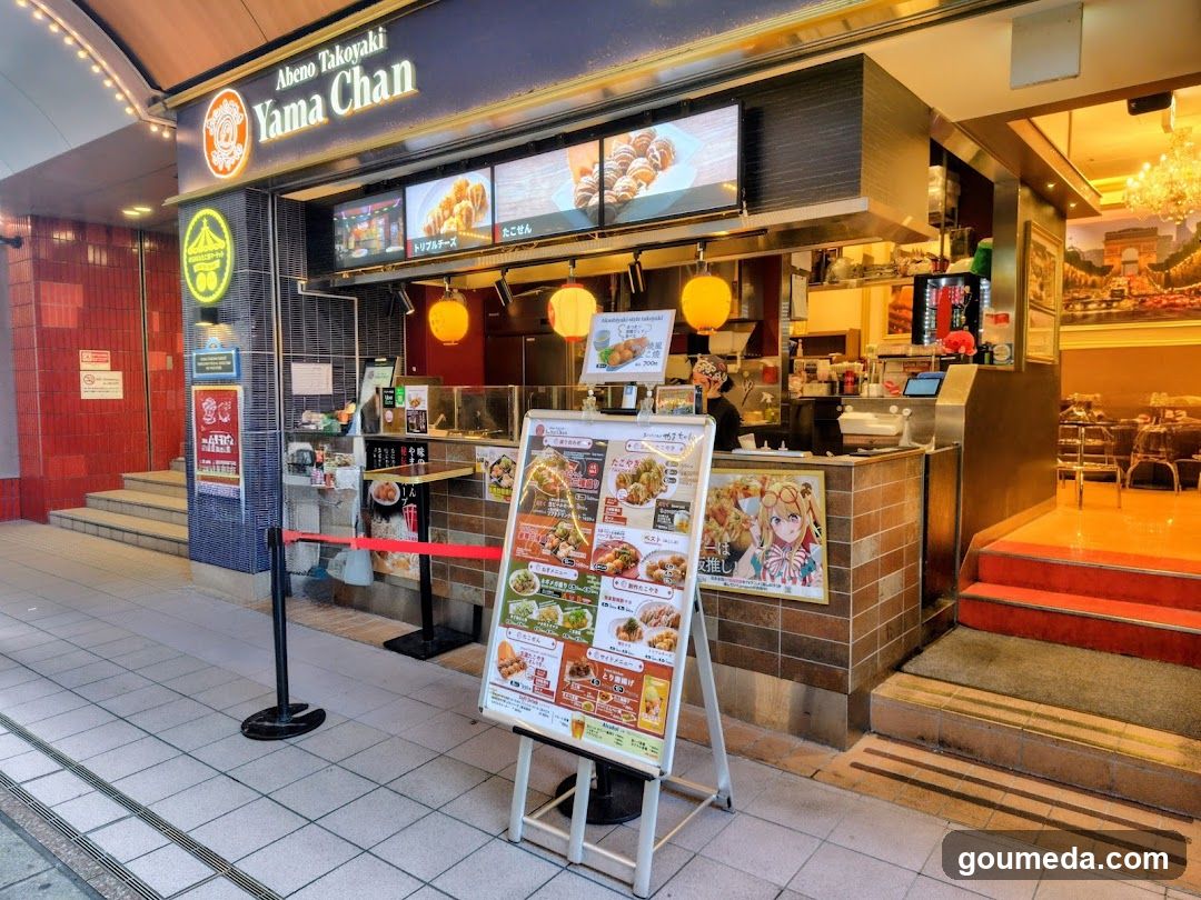 The image shows the exterior of a food stall named 'Yama Chan' with visible signage above the entrance. The shop features illuminated signs displaying menu items, including what appears to be takoyaki and other Japanese street food. A large menu board stands in front of the shop, showcasing various dishes with prices. The interior is partially visible through an open doorway, revealing warm lighting and seating areas.
