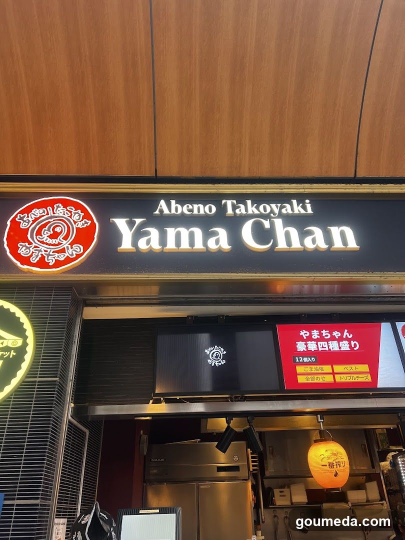 The image shows the storefront of a shop named 'Abeno Takoyaki Yama Chan'. The sign features a red circular logo with Japanese characters and the name in English. Below, there is a menu board displaying options like 'Yama-chan' with four types of toppings.