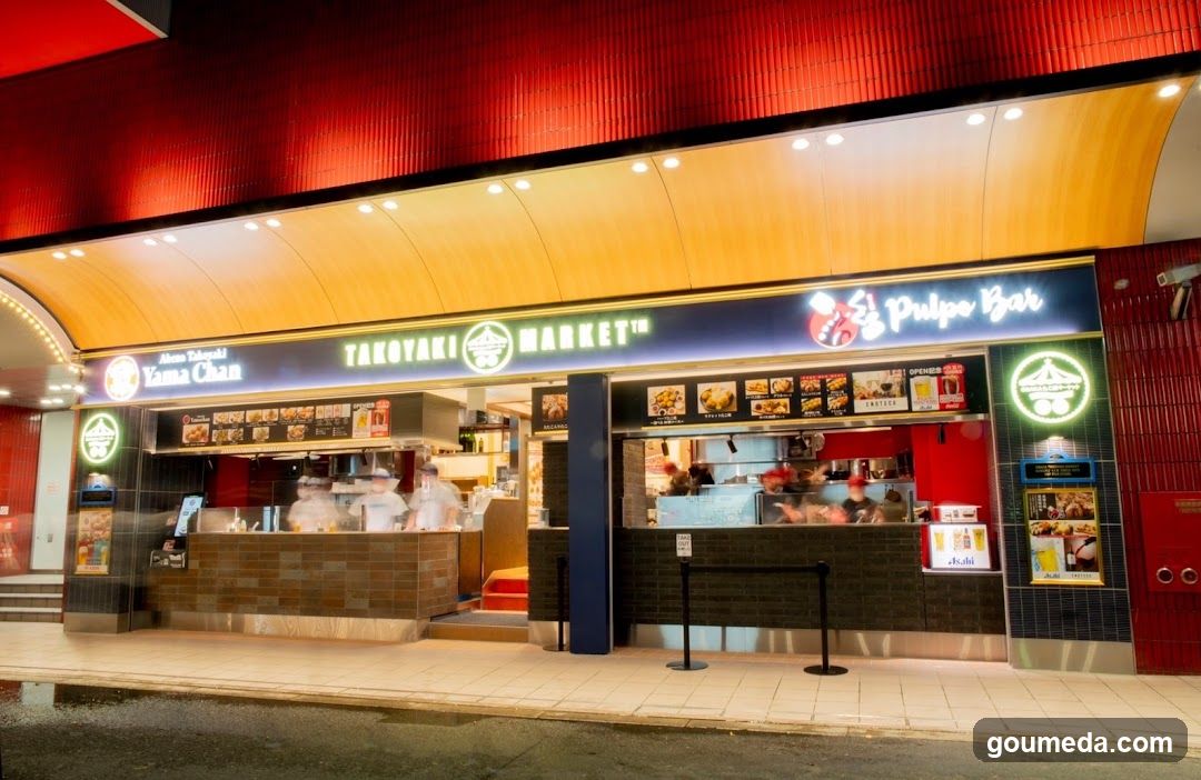 The image shows the exterior of a food court area with two visible stalls. On the left is 'Yama Chan Takoyaki', where chefs in white uniforms are preparing takoyaki on a wooden counter. To the right is 'El Pulpo Bar', featuring a glass window display showing various seafood items, likely octopus-based dishes given its name. Both signs are illuminated above their respective counters against a dark blue background. The ceiling above has warm lighting and red accents.