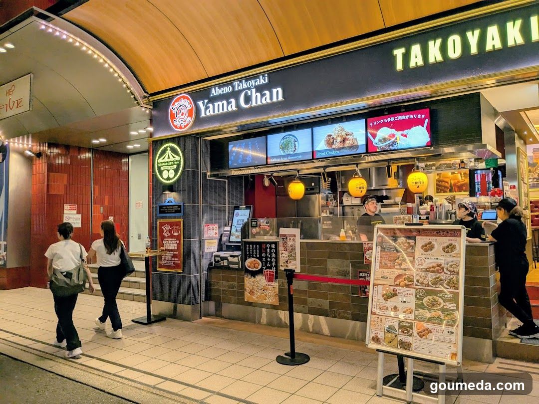 The image shows the storefront of Yama Chan, a takoyaki shop with illuminated signage reading 'Abeno Takoyaki Yama Chan' and 'TAKOYAKI'. The entrance features a tiled facade with hanging yellow lanterns. A menu board is visible outside, displaying various food items with prices. Staff members are seen inside the shop, preparing food behind the counter. Two customers are walking past the shop.