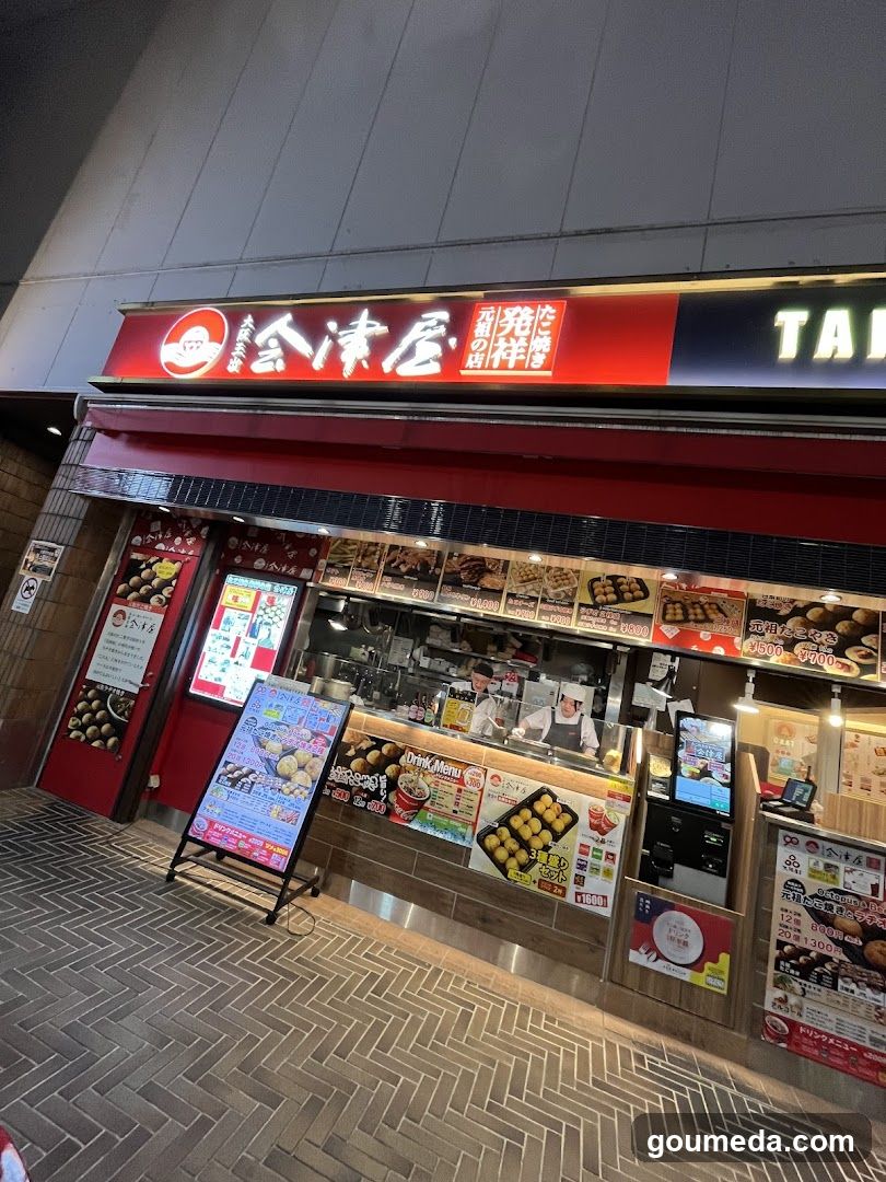 A brightly lit storefront with a red and black signboard displaying 'Tama Doya' in Japanese characters. The shop features a glass counter where staff are preparing food, visible behind the counter. Various menu boards and posters advertise different food items, including what appears to be takoyaki or similar street food. The entrance is accessible via a tiled walkway.