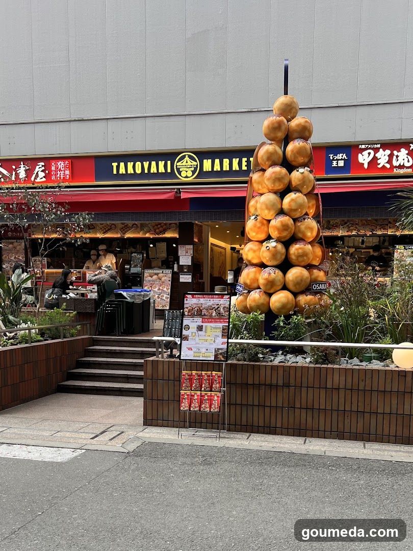 The image shows the exterior of a shop called 'TAKOYAKI MARKET' with visible signage on the building facade. A large decorative display made of takoyaki balls stands prominently in front, surrounded by greenery and planters. There are stairs leading up to the entrance, where staff can be seen inside. Menu boards and promotional materials are placed near the entrance.