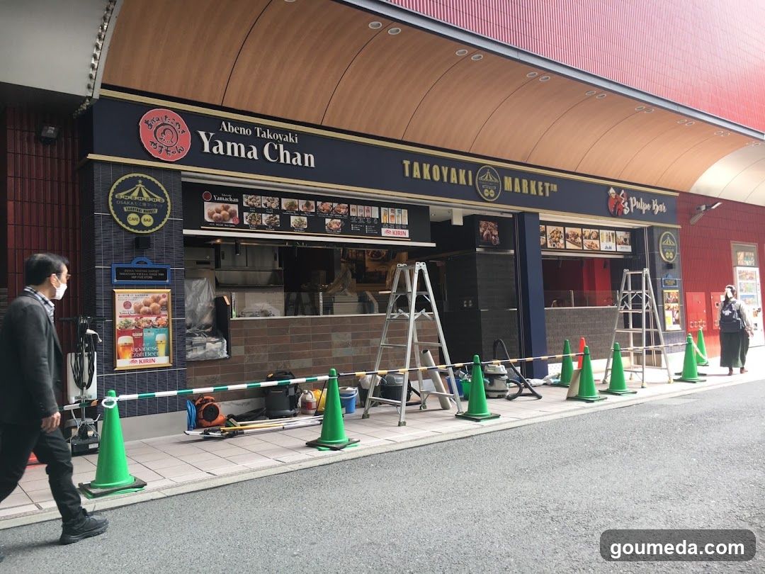 A storefront of Yama Chan Takoyaki Market, featuring a blue and red facade with visible signage. The shop has a menu board displaying various takoyaki items. Construction ladders and green traffic cones are placed in front of the entrance.