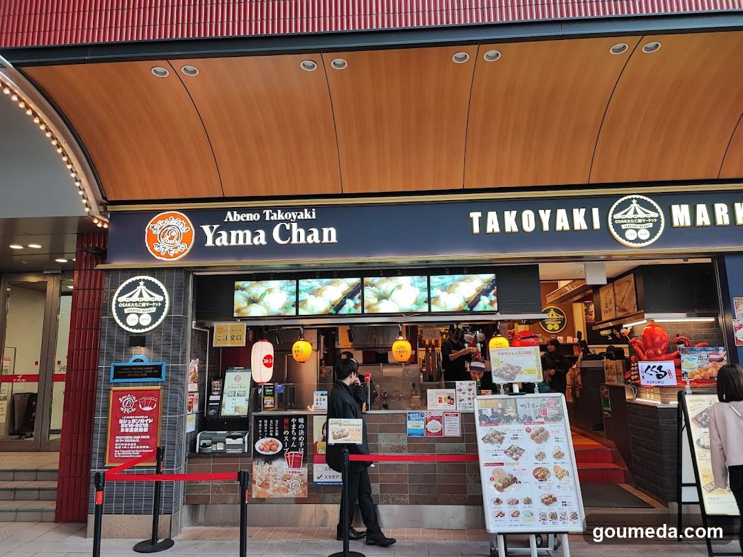 The image shows the storefront of Abeno Takoyaki Yama Chan, a takoyaki shop with a wooden facade and illuminated signage. The entrance is marked by red ropes and a queue system. Inside, staff are preparing food behind a glass counter. Menu boards display various takoyaki options.