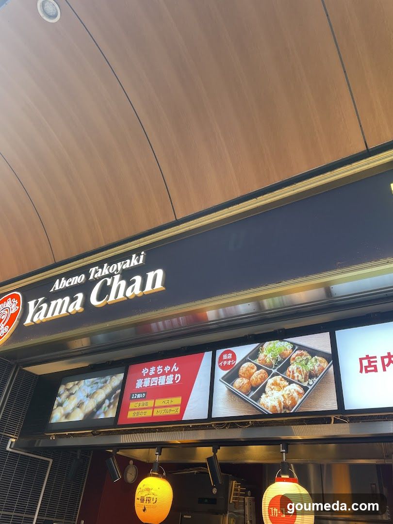 The image shows the storefront of a Yama Chan shop, featuring a prominent sign with 'Abeno Takoyaki' and 'Yama Chan' written on it. Below the sign, there are illuminated displays showcasing various takoyaki products, including different flavors and sizes. The shop has a modern design with wooden paneling above the signage.
