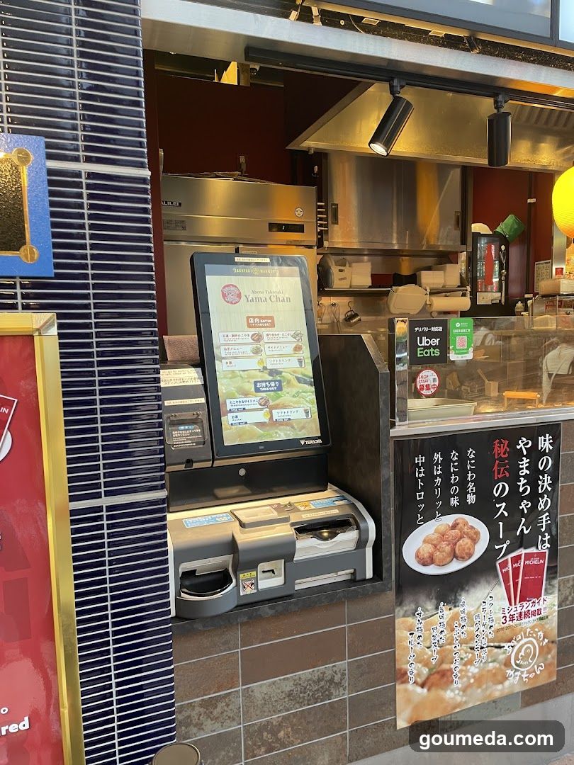 A shop front featuring a digital menu board displaying food items and prices, with visible signage including 'Uber Eats' and Japanese text advertising 'secret soup'. The interior shows stainless steel kitchen equipment and cooking areas.