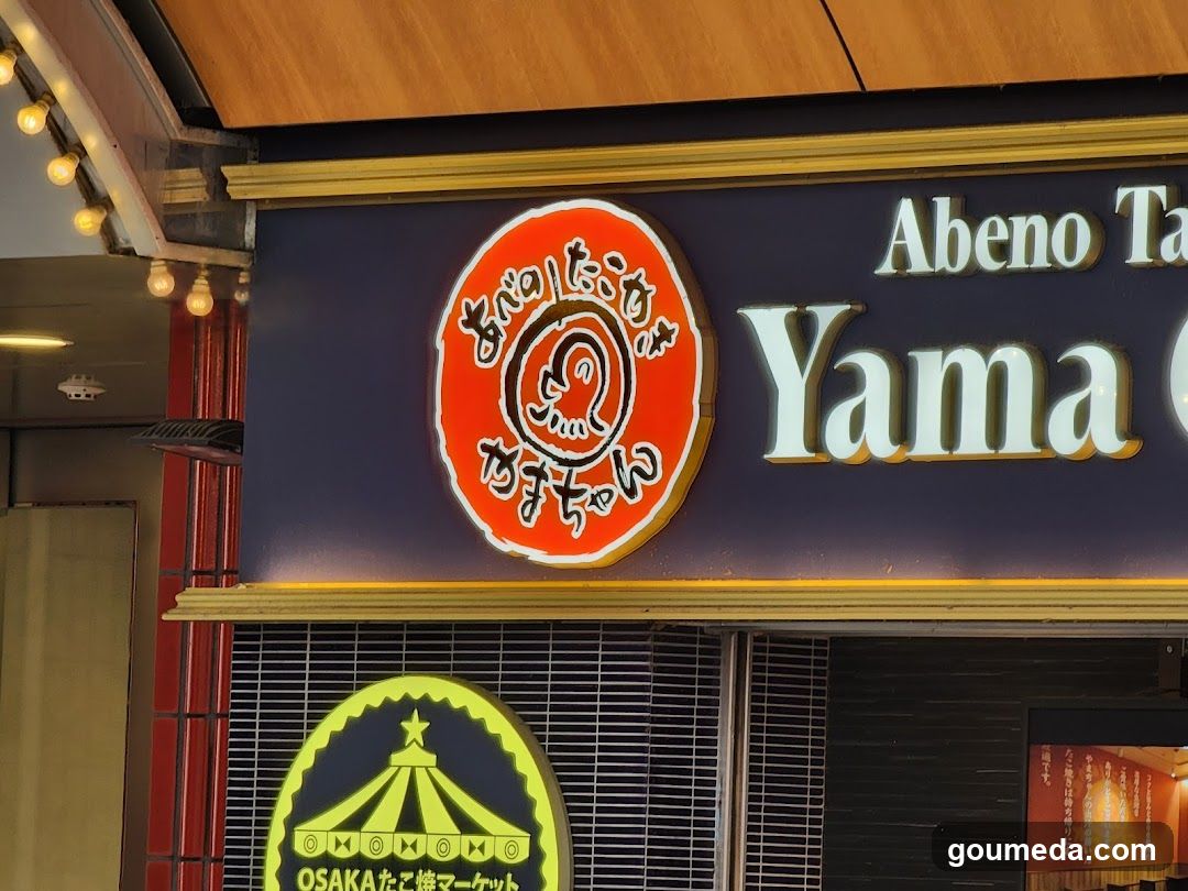 A shop front with a dark blue facade featuring illuminated signage. A circular red logo with Japanese characters and a stylized face is visible on the left. To its right, white lettering reads 'Abeno Ta' above larger text starting with 'Yama'. Below, a yellow emblem displays a tent-like structure and partial Japanese text. The entrance has metal grilles covering windows or doors.