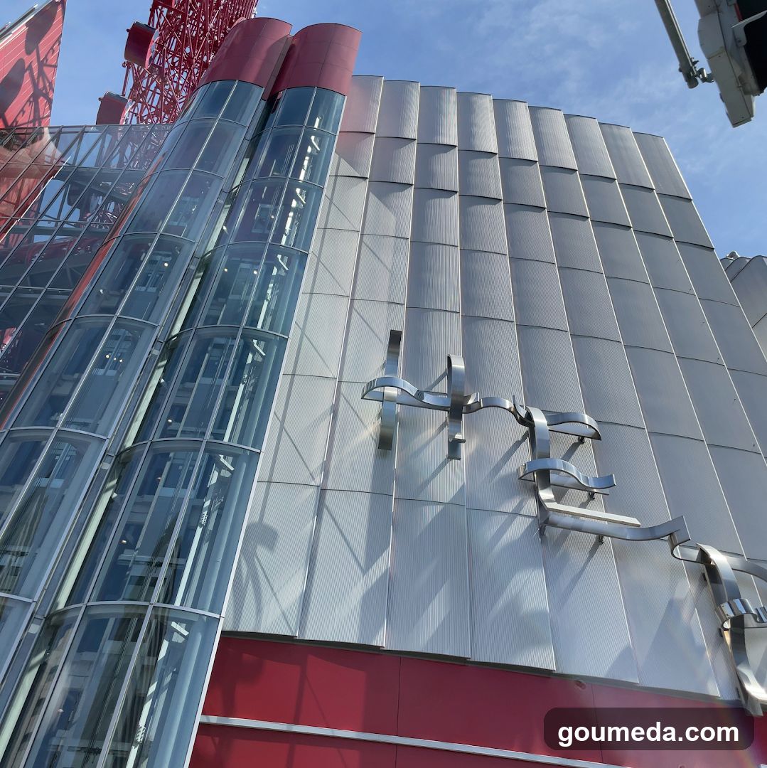 The image shows the exterior of a modern building featuring glass-enclosed elevators on the left side, metallic cladding panels covering most of the facade, and red accents at the bottom. A large stylized metal sculpture or sign is mounted on the wall. The sky is clear blue in the background.