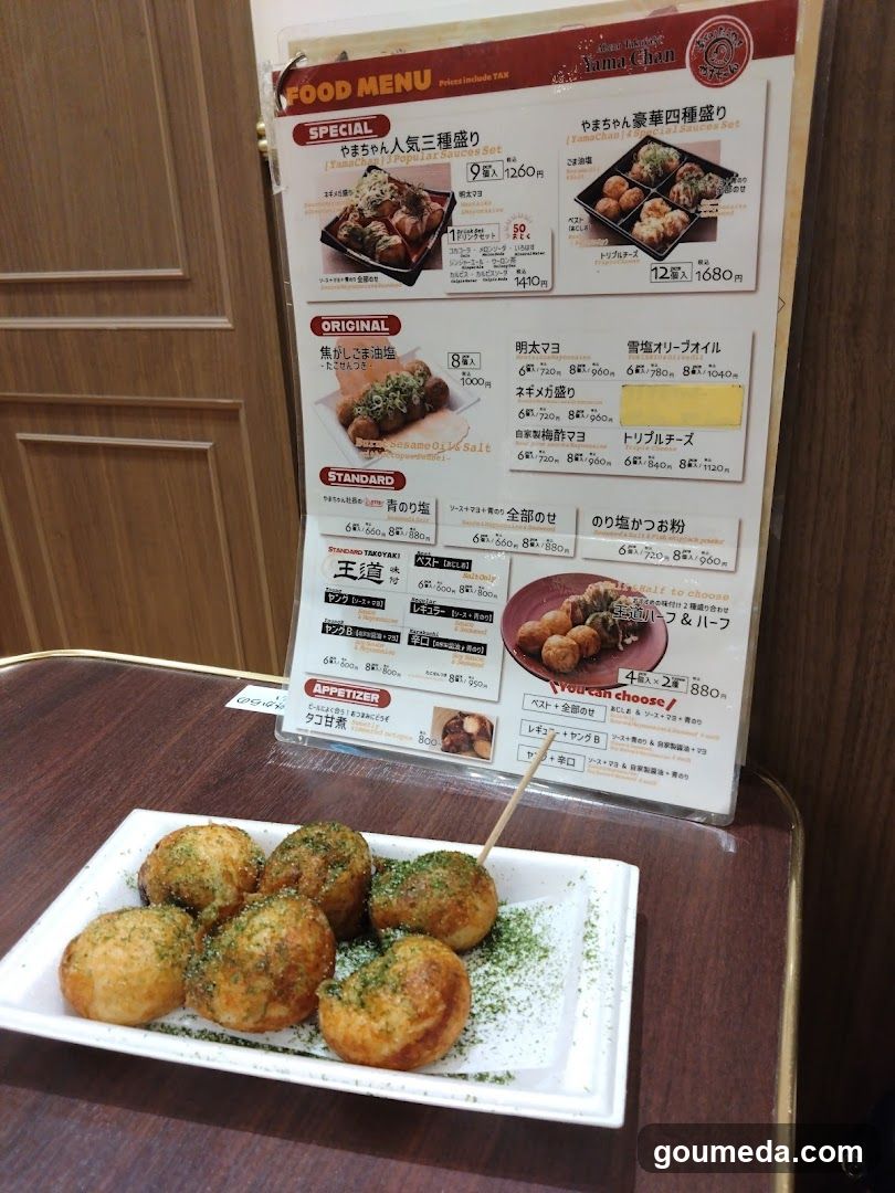 A plate of green-topped fried balls (otaku dango) sits on a table in front of a vertical menu board displaying Japanese food items and prices.