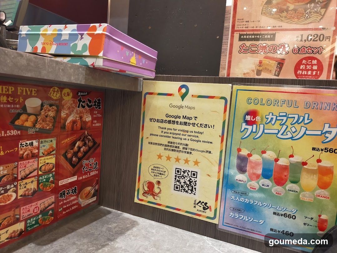 Interior shot of a Tokyo Disney Store counter area featuring promotional materials and merchandise. Visible items include colorful metal tins with 'TOKYO DISNEY STORE' branding, menu boards displaying takoyaki sets and drinks like Karafuru Cream Soda, and Google Maps review QR codes.