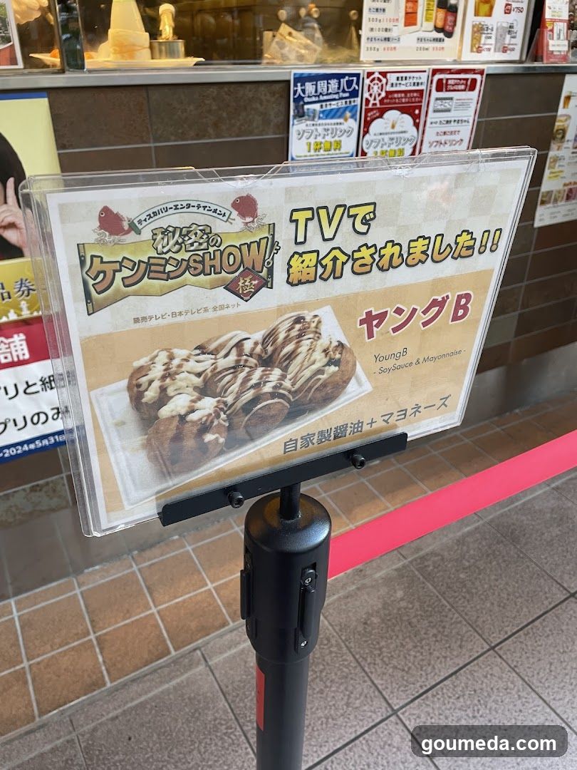 A clear acrylic signboard displaying a promotional advertisement featuring 'Keenmin Show' and 'Young B', showcasing a dessert with chocolate drizzle and mayonnaise sauce, alongside Japanese text promoting TV appearances.