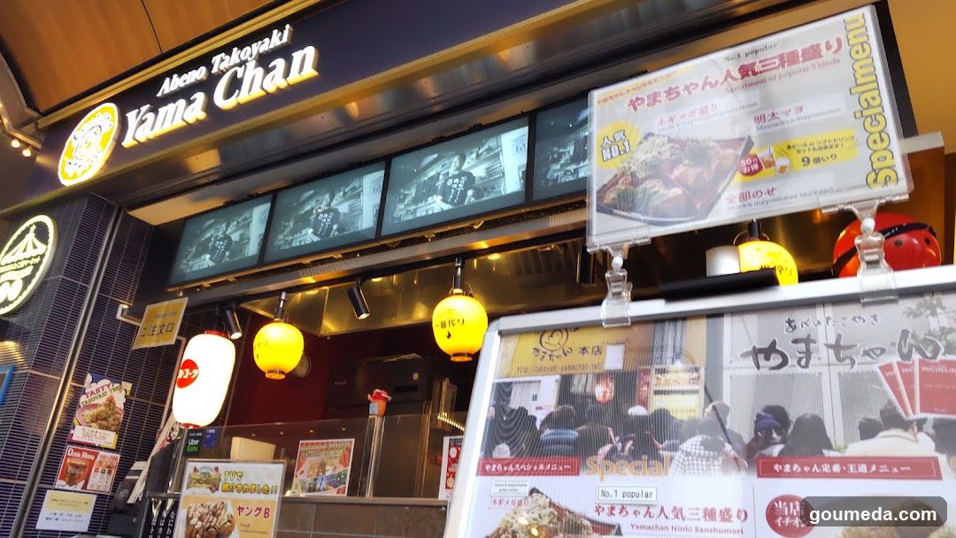 The image shows the exterior of a shop named Yama Chan, featuring illuminated signage with Japanese and English text. There are multiple screens displaying food items above the entrance. Yellow lanterns hang from the ceiling inside, adding to the ambiance. A menu board is visible on the right side, showcasing various dishes.