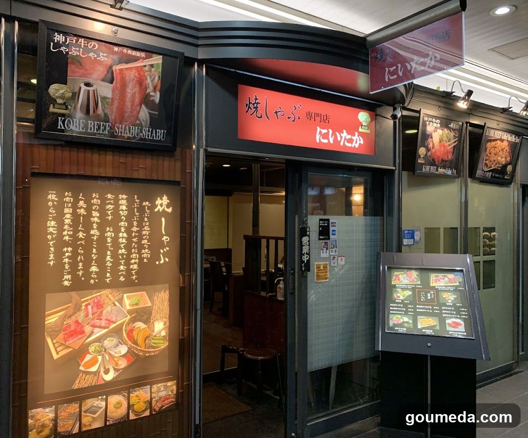 The image shows the storefront of a Japanese shabu-shabu restaurant named 'Nito Kake' (にいたか). The entrance features glass doors with signage above indicating it is a specialty shop. To the left, there are posters advertising beef cuts and menu items including grilled skewers and hot pot dishes. A digital menu board stands outside displaying various food options with prices in yen.