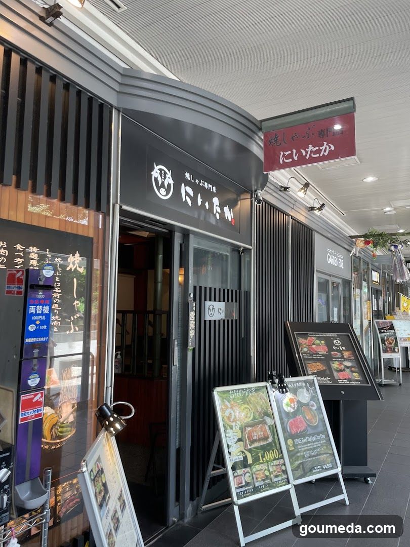 A modern storefront with black and white vertical slats, featuring a sign reading 'にいたか' (Nikitaka) above the entrance. A hanging red banner displays Japanese text about grilled fish. Outside menus are placed on stands near the door.